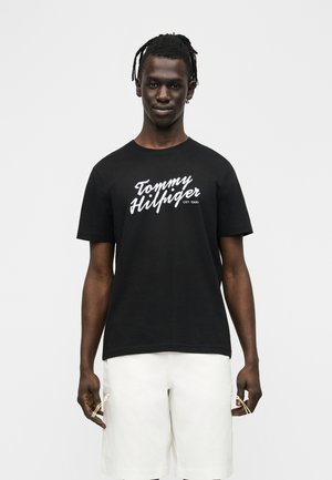 Moški s spetimi lasmi, oblečen v črno majico "Tommy Hilfiger" in bele kratke hlače, stoji na preprostem svetlo sivem ozadju.