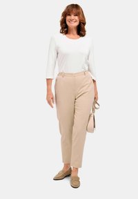 Beige maßgeschneiderte Hose mit hoher Taille, weißes Langarmhemd und hellbraune Slipper. Als Accessoires dient eine kleine hellfarbige Handtasche.