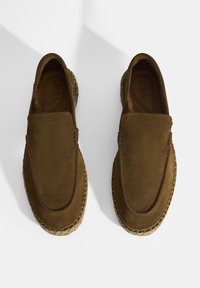 Bruine suède instappers met een ronde neus en geperst jute detail langs de zool. Gekozen textuur en minimale stikselaccenten.