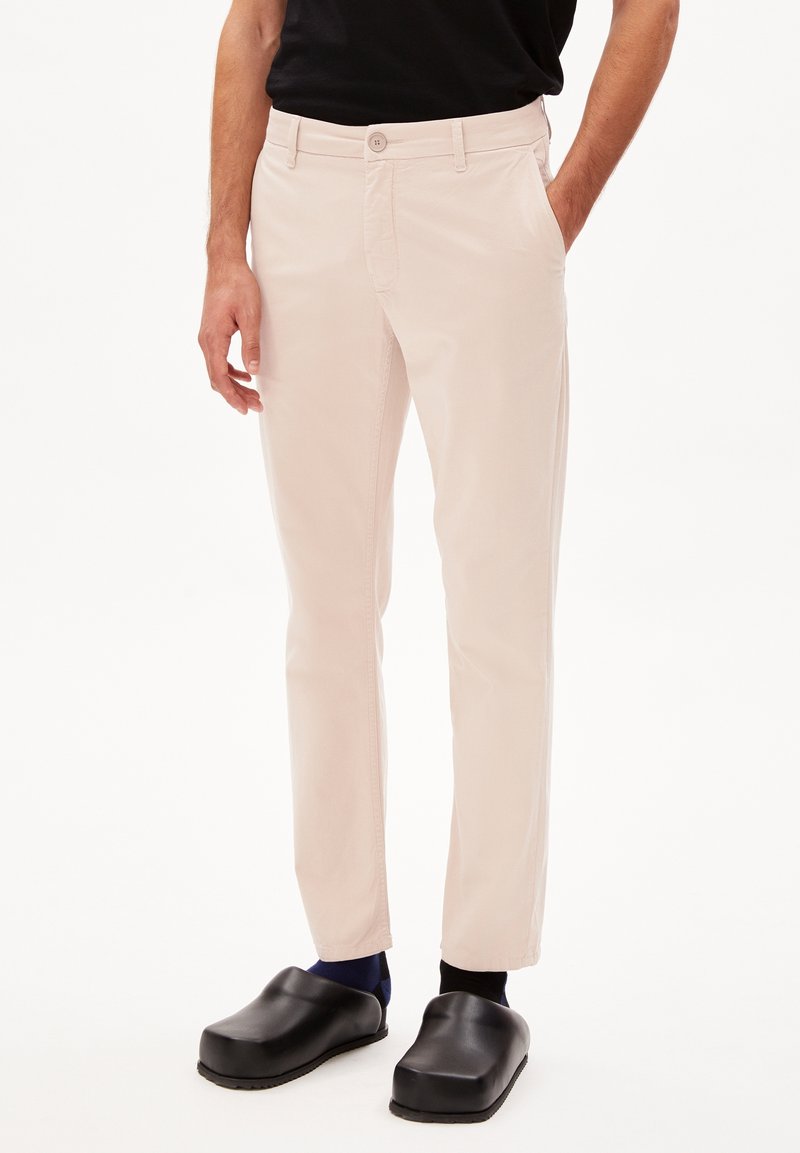 Světle růžové chinos kalhoty s knoflíkovým zapínáním a diagonálními kapsami, kombinované s černými dřeváky a modrými ponožkami. Materiál má hladkou texturu.