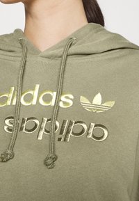 Green hoodie with shiny gold Adidas logo and trefoil emblem on chest, worn by a person with dark hair pulled back.