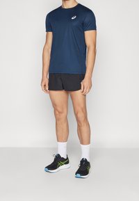 Pánský sportovní outfit skládající se z navy modrého trička s krátkým rukávem, černých šortek a černých běžeckých bot s zelenými a modrými akcenty.