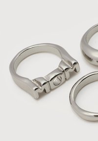 Τρία λαμπερά ασημένια μεταλλικά δαχτυλίδια, το ένα με ανάγλυφα γράμματα που σχηματίζουν τη λέξη "MOM" γύρω από το δαχτυλίδι, πάνω σε λευκή επιφάνεια.