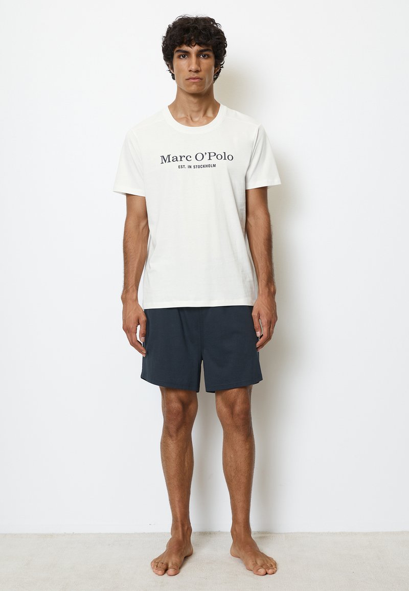 Vit bomull T-shirt med svart tryck "Marc O'Polo EST. I STOCKHOLM," kombinerad med mörkblå shorts, bärs av en stående manlig modell.