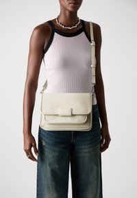Krémeově zbarvená kožená crossbody taška s texturovaným povrchem, klopovým uzávěrem a odnímatelným popruhem. Nositelná s upnutým tílkem a širokými džíny.