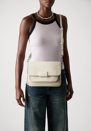 Sac en cuir couleur crème à bandoulière avec une surface texturée, fermeture à rabat et bandoulière amovible. Porté avec un débardeur ajusté et un jean évasé.