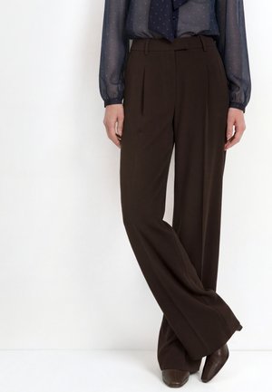 Vrouw draagt hoge taille, wijde bruine broek, doorzichtige donkerblauwe blouse met lange mouwen en polkadots, en bruine puntige schoenen.