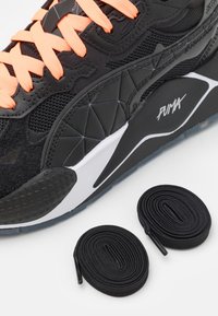 Svart atletisk joggesko med oransje snørebånd, med en teksturert mesh-overdel, geometriske designelementer, og to svarte snørebånd ved siden av.