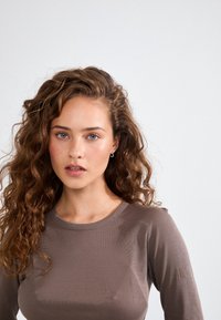 Pikendusega, ümmarguse kaelusega tops pehmest, ribakujulisest pruunist kangast. Omab diskreetset logo rinnatükil. Mudelil on looduslikud, lahtised lokid.