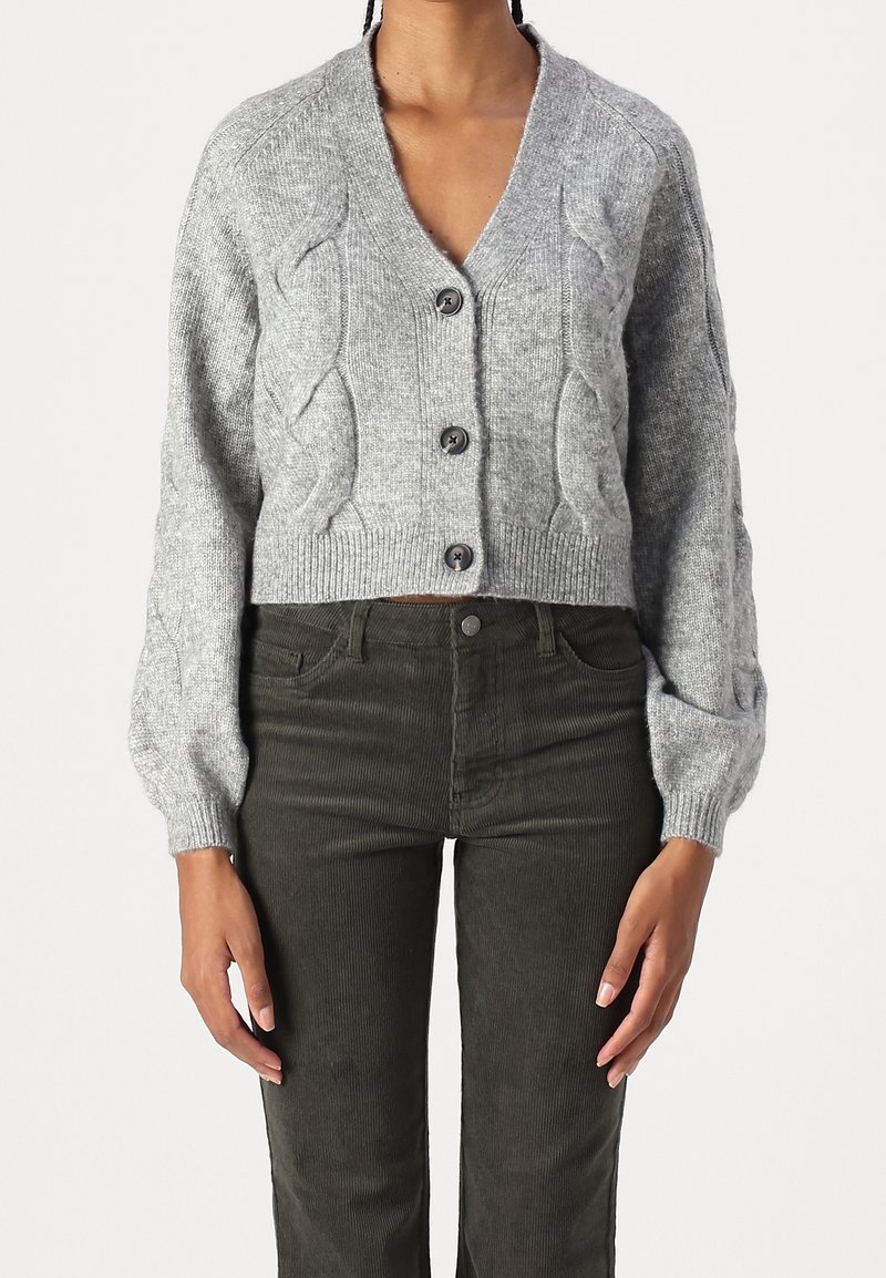 Cardigan gris en tricot avec motif torsadé et boutons, porté avec un pantalon en velours côtelé vert olive foncé.
