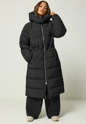 Femme portant un long manteau matelassé noir avec un col montant, un pantalon large noir et des baskets blanches, debout avec une main près de son col.