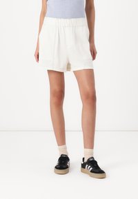 Shorts en coton blanc avec une taille élastique, des poches latérales, une coupe ample et un ourlet roulé. Portés avec des chaussures noires et des chaussettes beiges.