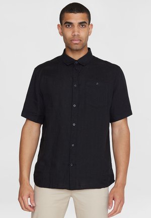 Mann trägt ein kurzärmeliges schwarzes Hemd mit Knöpfen und Kragen sowie beige Hosen vor einem schlichten Hintergrund.