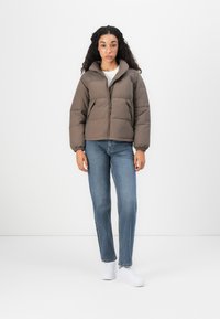 Giubbotto imbottito marrone con colletto alto, chiusura con zip e polsini elastici, abbinato a jeans blu e sneakers bianche su uno sfondo neutro.