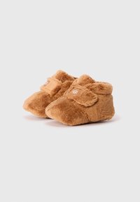 Chaussons en peluche marron avec une texture douce, une sangle Velcro, une forme arrondie et une petite étiquette de logo sur le côté, conçus pour le confort.