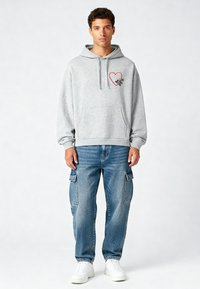 Felpa grigia con cappuccio, decorata con una grafica a cuore rosso e dettagli neri, abbinata a pantaloni cargo blu e sneakers bianche.