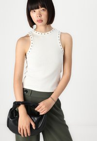 Witte geribde mouwloze top met studaccenten bij de halslijn, gecombineerd met donker groene high-waisted broek en een zwarte handtas.