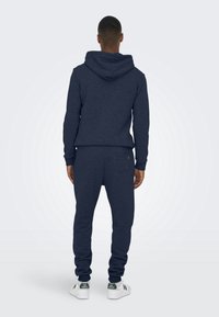 Marineblaue Kapuze und passende Jogginghosen aus weichem, strukturiertem Stoff. Verfügt über eine Kapuze und elastische Bündchen. Das Model trägt weiße Sneaker mit grünen Akzenten.