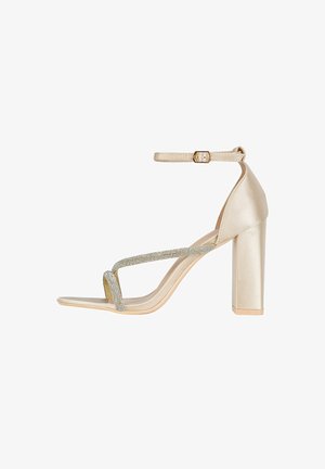 Sandale à talons hauts beige avec une finition satinée, bride argentée ornée, talon bloc, bout ouvert et bride de cheville avec une boucle dorée.