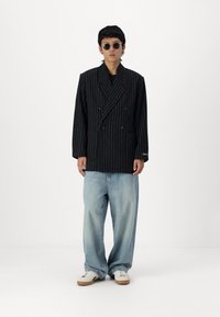 Double-breasted black pinstripe blazer with wide lapels, paired with light blue loose-fit jeans and white sneakers. Accessories include round sunglasses.