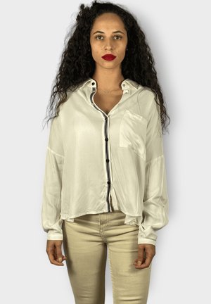 Camisa de botones de seda blanca con una franja negra a lo largo de la placket, un bolsillo en el pecho y mangas largas, usada con pantalones de color claro.