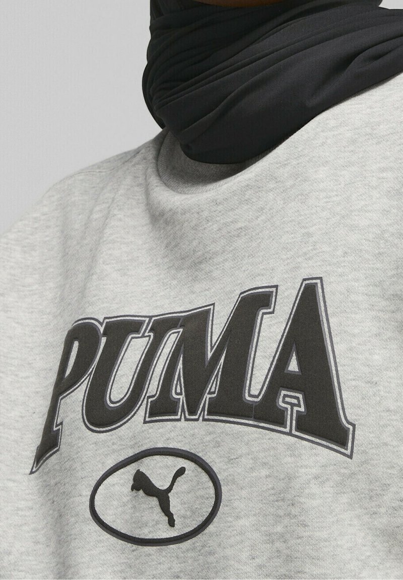 Felpa grigia con stampa e logo "PUMA" neri in un ovale. Il materiale sembra morbido e di texture, con un colletto rotondo. Accessorio nero visibile attorno al collo.