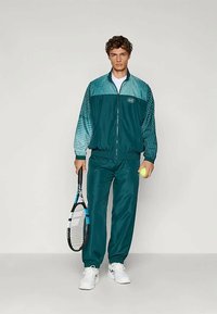 Tirkizni trenerka od glatkog materijala koja sadrži jaknu na zatvarač s gradijentom boja i uzorcima; uključuje tenisku reketa i lopticu.