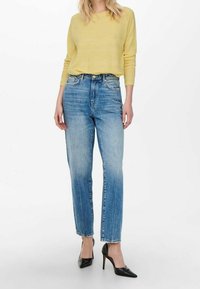 Pull texturé jaune clair, jean bleu taille haute avec des zones délavées, et escarpins noirs à bout pointu. Coupe droite avec une coupe décontractée.