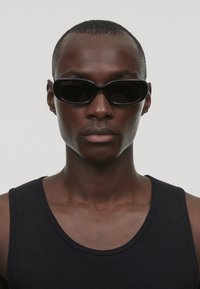 Ochelari de soare negri cu o formă elegantă, având o ramă subțire și lentile întunecate, purtați de o persoană în tricou fără mâneci negru, evidențiind un profil facial clar.