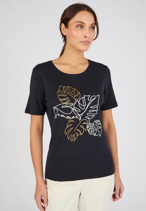 Mujer con cabello castaño con una camiseta negra de manga corta con contornos de hojas tropicales amarillas y blancas y la palabra "verano", que lleva pantalones claros.