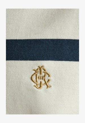 Cremefarbener Strickstoff mit einem einzelnen dunkelblauen horizontalen Streifen und einem kleinen beige bestickten Monogramm unterhalb des Streifens.