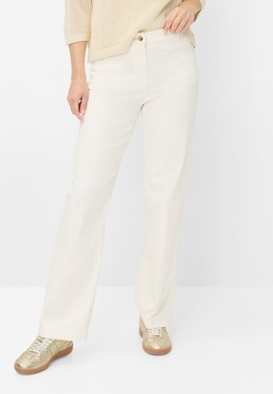 Kvinde iført off-white bukser med lige ben, beige striktrøje og guld sneakers med hvide snørebånd, stående mod en hvid baggrund.