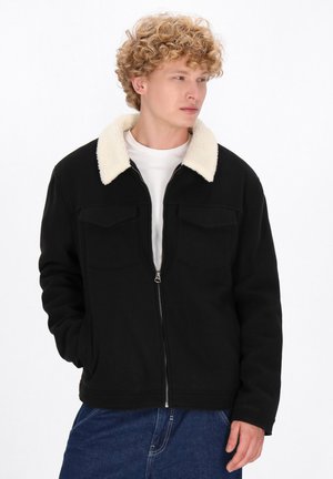 Ung mand med krøllet hår iført en sort jakke med lynlås og hvid fleece-krave samt blå jeans, der ser til siden med en hånd i lommen.