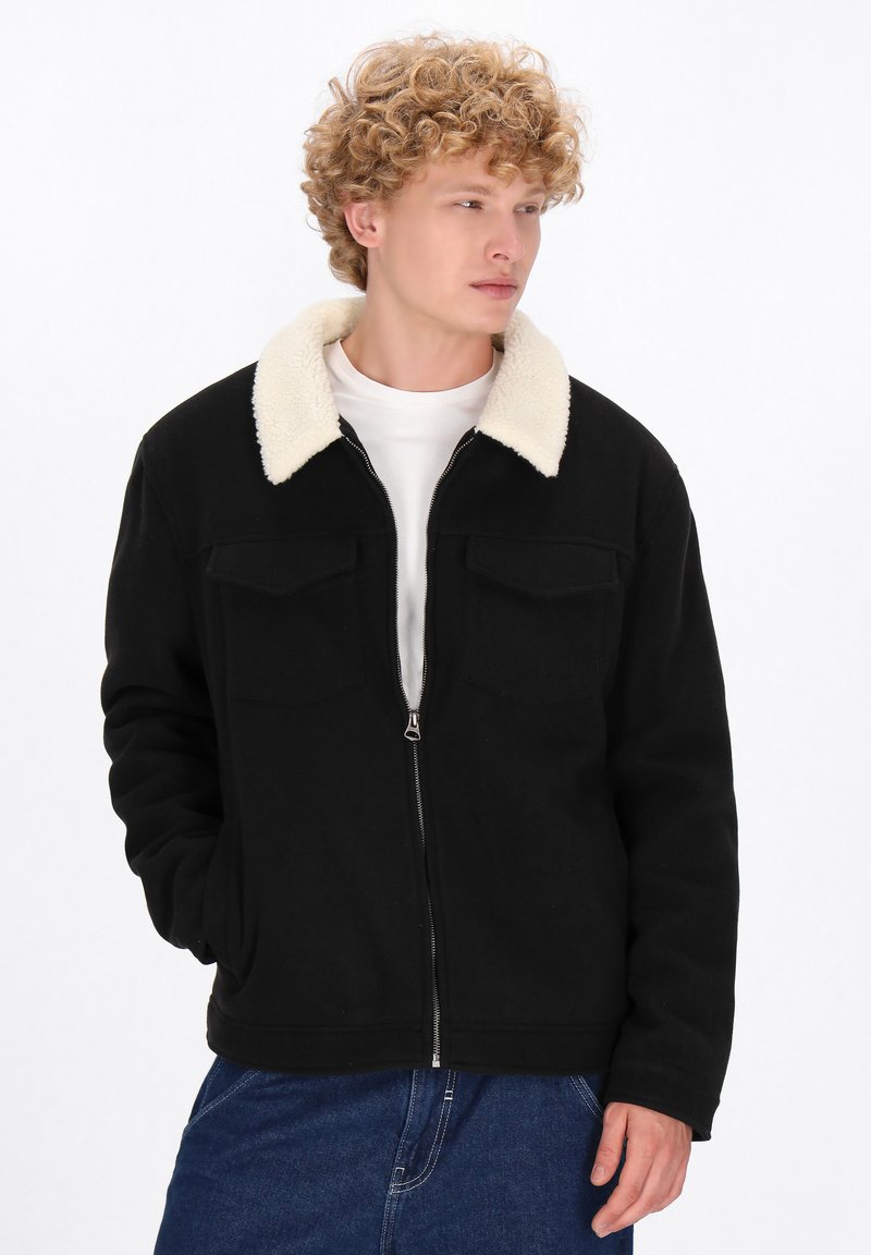 Giovane uomo con capelli ricci che indossa una giacca nera con zip e colletto in pile bianco e jeans blu, che guarda di lato con una mano in tasca.