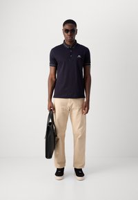 Marineblauw poloshirt met een drieknoopssluiting, contrasterende kraag en mouwstrepen, gecombineerd met beige broeken en zwarte sneakers, met een zwarte tas in de hand.