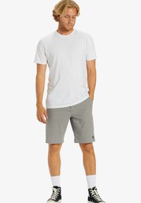 T-shirt en coton blanc associé à un short gris ; le short présente une texture lisse et une petite étiquette avec un logo sur le devant. Chaussures montantes noires.
