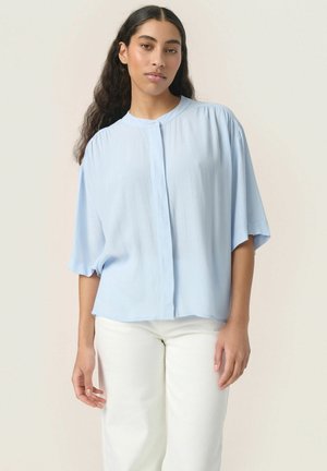 Blusa leggera azzurro chiaro a maniche corte con colletto rotondo, chiusura a bottoni e vestibilità ampia. Realizzata in tessuto morbido e leggero con una sottile trama.