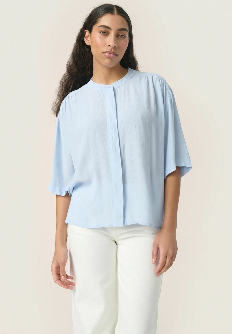 Blusa leggera azzurro chiaro a maniche corte con colletto rotondo, chiusura a bottoni e vestibilità ampia. Realizzata in tessuto morbido e leggero con una sottile trama.