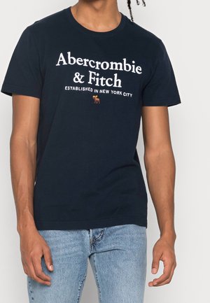 Osoba ubrana w granatową koszulkę Abercrombie & Fitch z białym napisem i małym logo łosia, w połączeniu z jasnoniebieskimi dżinsami.