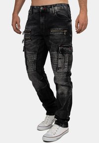 Black denim cargo pants featuring multiple zippered pockets, ribbed accents, and a tapered fit. Finished with white sneakers for contrast.