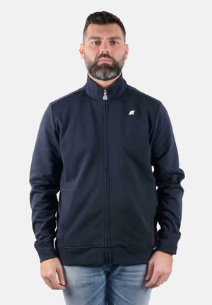 Giacca a zip blu navy con collo alto, cuciture strutturate e un piccolo logo. Il tessuto appare liscio ed elastico, adatto per l'abbigliamento attivo.