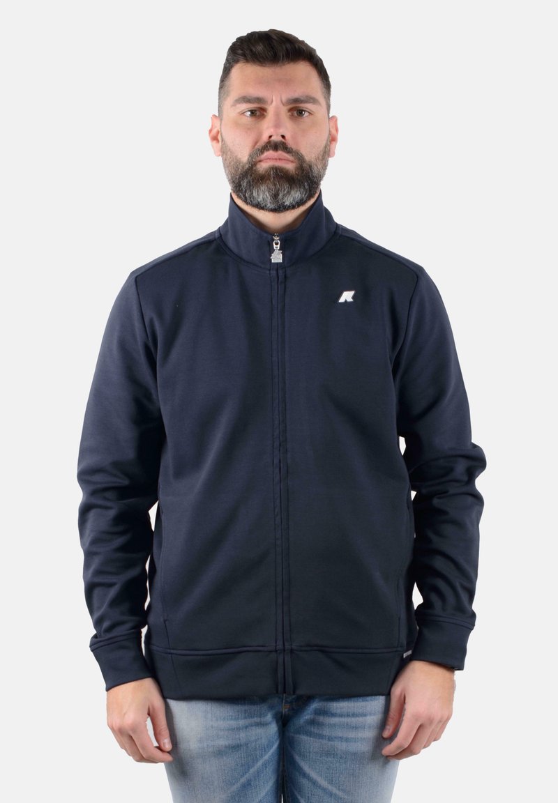 Giacca a zip blu navy con collo alto, cuciture strutturate e un piccolo logo. Il tessuto appare liscio ed elastico, adatto per l'abbigliamento attivo.