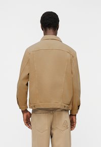 Άνδρας που φοράει μπεζ μπουφάν και παντελόνι που ταιριάζει με λουλουδάτη κεντητή λεπτομέρεια στην πίσω τσέπη, στέκεται με την πλάτη προς την κάμερα.