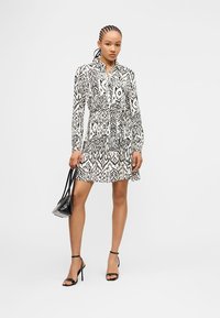 Γυναίκα με πλεκτά μαλλιά που φοράει μακρυμάνικο φόρεμα σε ασπρόμαυρο μοτίβο, μαύρες γόβες και κρατάει μια μαύρη τσάντα.