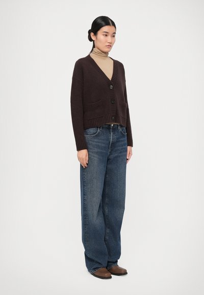 Dark brown cardigan with a v-neck, button closure, and ribbed texture. Paired with loose-fit blue jeans and brown slip-on shoes.