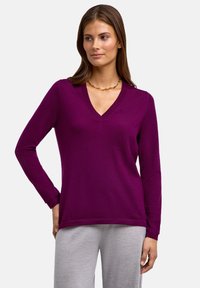 V-ausschnitt lila Pullover aus weichem Stoff mit langen Ärmeln, im l�ässigen Schnitt und mit subtilen Saumdetails. Kombiniert mit grauen Hosen.