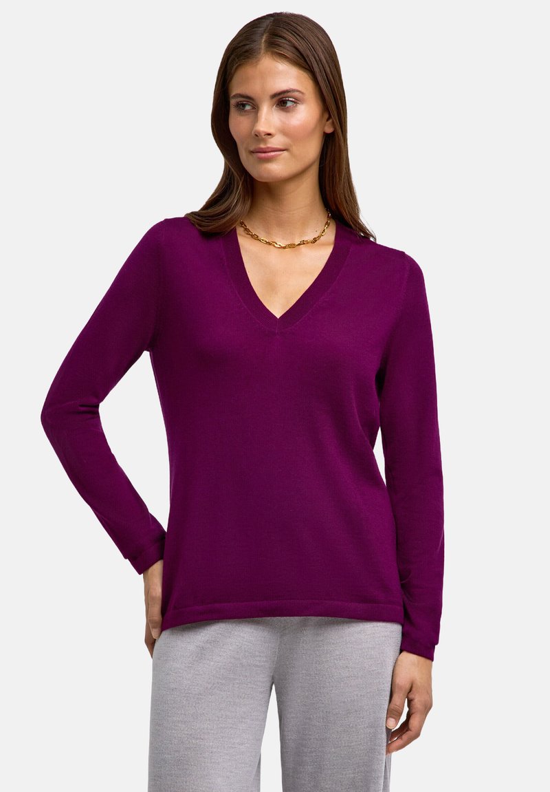 V-ausschnitt lila Pullover aus weichem Stoff mit langen Ärmeln, im lässigen Schnitt und mit subtilen Saumdetails. Kombiniert mit grauen Hosen.