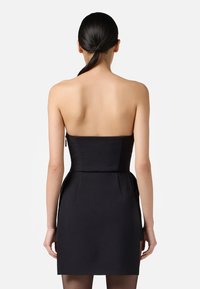 Donna con coda di cavallo nera che indossa un mini abito nero senza spalline, di spalle contro uno sfondo bianco uniforme.