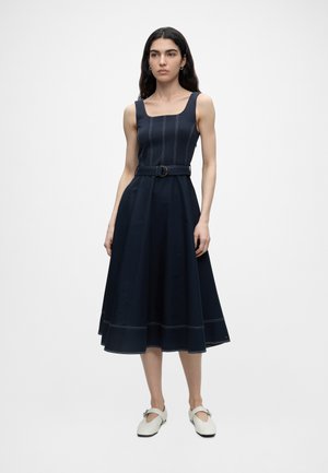 Žena v nosenej tmavomodrej midi šaty bez rukávov s opaskom a bielymi topánkami so sponou, stojaca na jednotnú bielu pozadí.