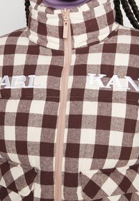 Kurtka puchowa w kratę w kolorze brązowym i beżowym z wysokim kołnierzem. Posiada zamek z przodu, haftowane logo "KARL KAN" oraz kieszenie na piersi.